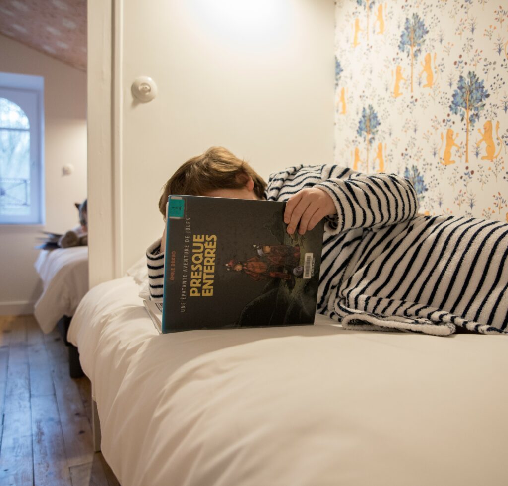 Chambre individuelle avec lit simple. Un enfant est allongé sur le lit entrain de lire une bd.